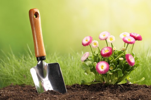 A close-up view of a garden bed featuring a freshly dug patch of dark soil with a small gardening trowel inserted into the ground in the foreground, which has a wooden handle and metal blade. To the right of the trowel, there is a cluster of pink and white daisy-like flowers with green foliage, suggesting a cultivated flower bed. The background consists of well-maintained, vibrant green grass that extends across the image, creating a lush outdoor environment typical of residential gardens in Coney Hall. Lighting is bright and natural, indicating a sunny day with a softly blurred background that emphasizes the garden elements in the foreground. This scene captures a moment of outdoor gardening activity, aligning with professional gardening services such as those offered in the Coney Hall area by Gardening Services Coney Hall, focusing on planting, lawn care, and garden maintenance.
