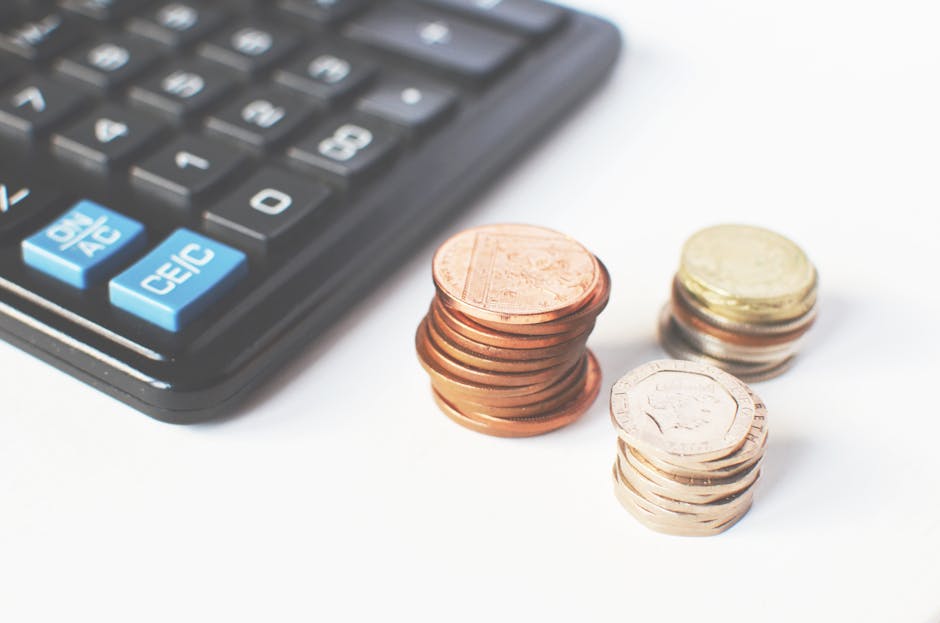 Close-up of a black calculator with light blue and white buttons, positioned next to several stacks of coins, including copper penny coins, a small pile of stacked euro coins, and a few loose coins, a